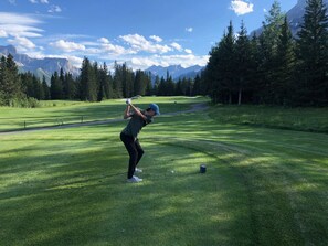 Una persona juega al golf en un campo bien cuidado, con cielo despejado y montañas al fondo.