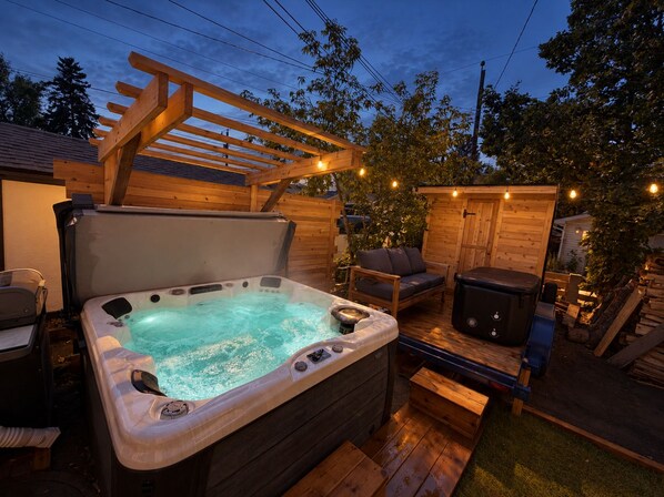 A hot tub with a wooden deck and outdoor furniture, surrounded by trees and a wooden fence.