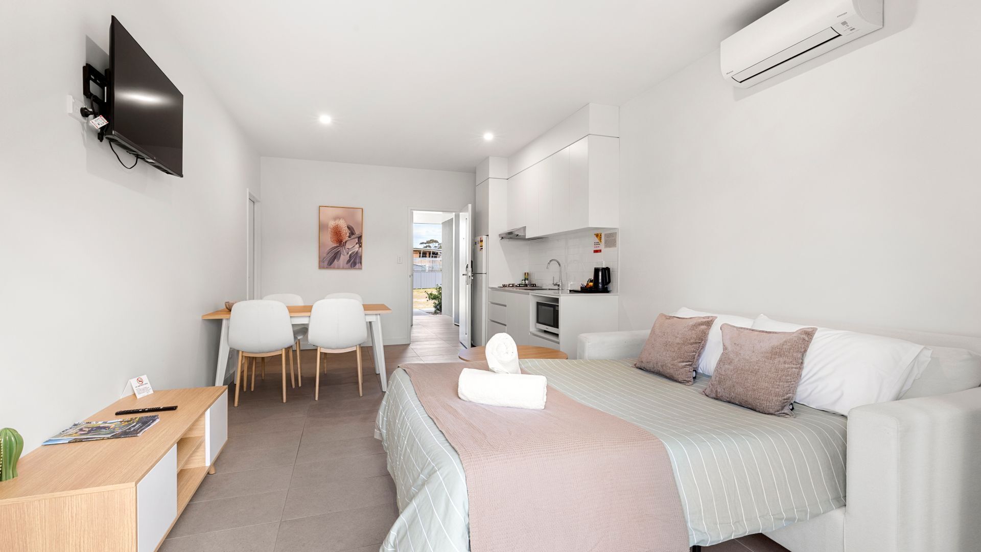 A modern, well-lit room with a bed, a desk, a chair, and a kitchenette.