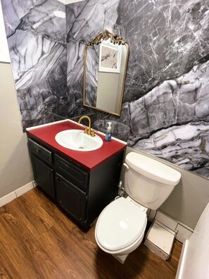 Een badkamer met een marmeren wand, een rode wastafelmeubel, een gouden kraan, een wit toilet en een spiegel met een versierd frame.