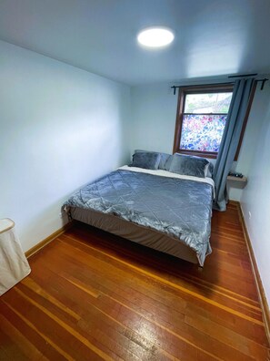 A small, well-lit bedroom with a single bed, a window with a view, and a wooden floor.