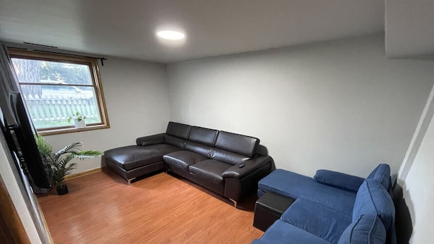 A living room with a brown leather sectional sofa, a blue sofa, and a small table. There is a window with a view of a fence and trees.