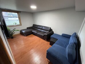 Een woonkamer met een bruine leren hoekbank, een blauwe bank en een kleine tafel. Er is een raam met uitzicht op een hek en bomen.