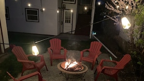 A fire pit surrounded by red Adirondack chairs in a backyard with string lights.