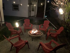 A fire pit surrounded by red Adirondack chairs in a backyard with string lights.