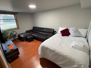 Een slaapkamer met een bed, een zwarte bank, een kleine tafel met een plant en een raam met uitzicht op een witte schutting.