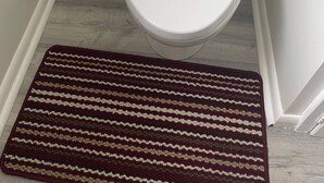 A toilet with a white lid and tank, a white trash bin, and a brown striped rug on a wooden floor. - Welcome to the Cabernet Escape– A Bold, Wine-Inspired Urban Escape (Calgary)
