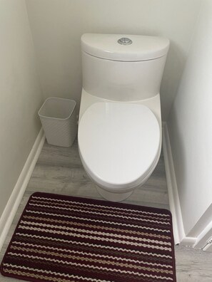 A toilet with a white lid and tank, a white trash bin, and a brown striped rug on a wooden floor.