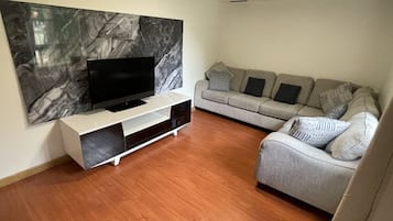 A living room with a flat-screen TV on a white stand, a grey sofa with patterned cushions, and a marble wall.