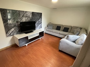 A living room with a flat-screen TV on a white stand, a grey sofa with patterned cushions, and a marble wall.