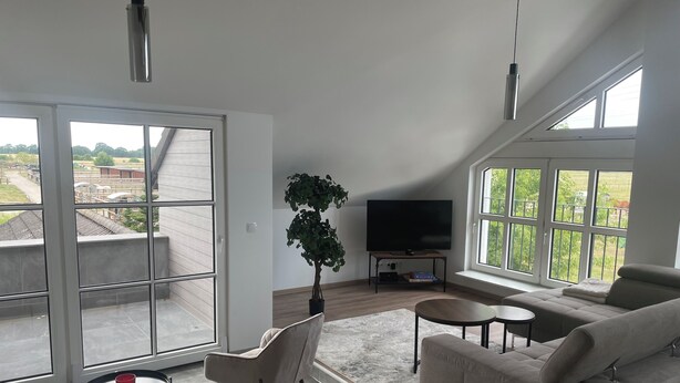 Una sala de estar moderna con un amplio ventanal corredera, un televisor de pantalla plana, un sofá color beige, una mesita redonda y una planta en maceta.
