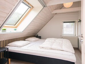 Une chambre avec un plafond en pente, un lit avec une literie blanche, une petite table et une fenêtre donnant sur un immeuble.