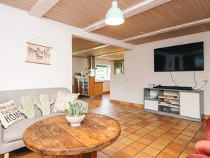 A living room with a wooden ceiling, a round coffee table, a sofa with cushions, a television on the wall, and a kitchen area in the background. - 4 Star Holiday Home in Henne (Henne)