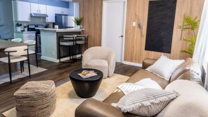 A modern living room with a sofa, ottoman, and coffee table. A kitchen with white cabinets and a dining area with chairs are visible in the background. - Cozy 3b2b Duplex Home in South Austin (Austin)
