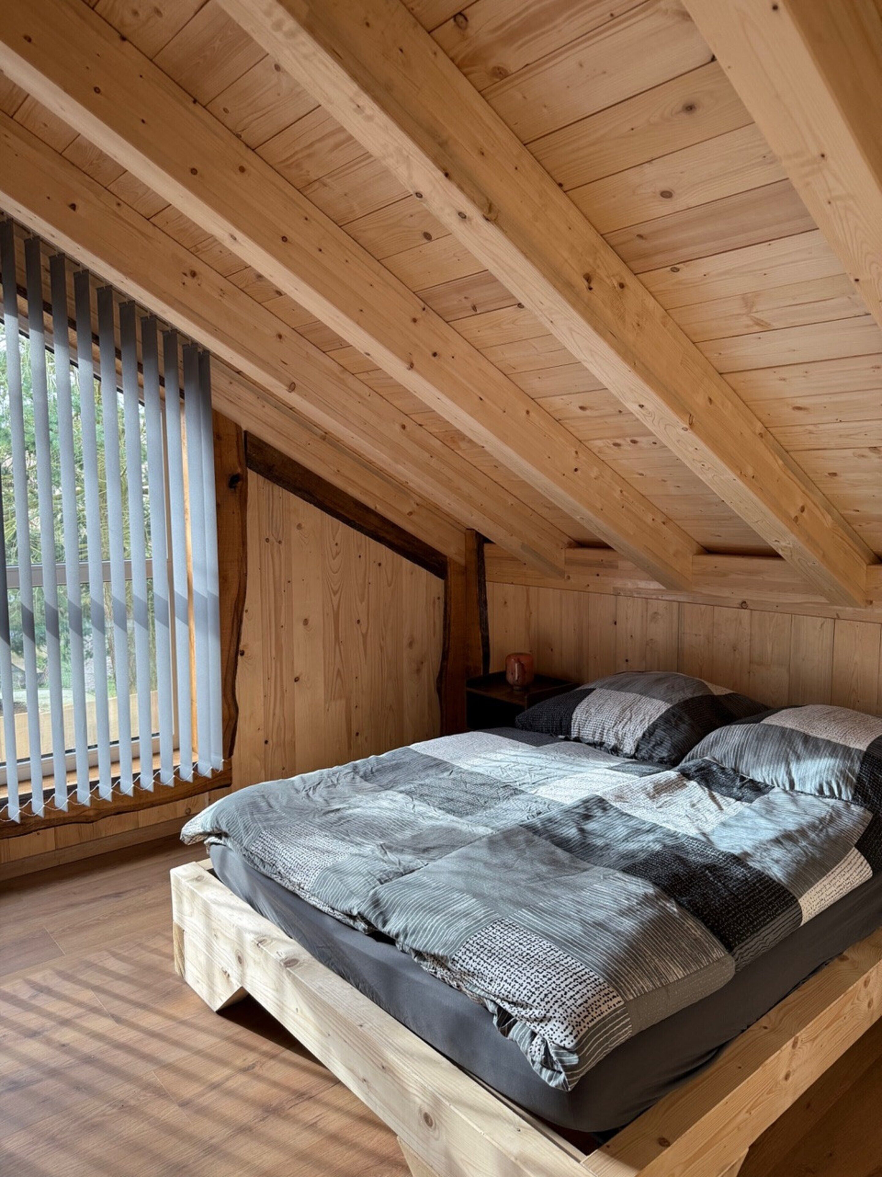 Une chambre à coucher aménagée dans les combles, avec un plafond en pente, un lit recouvert d’une literie grise et une fenêtre dotée de stores verticaux.