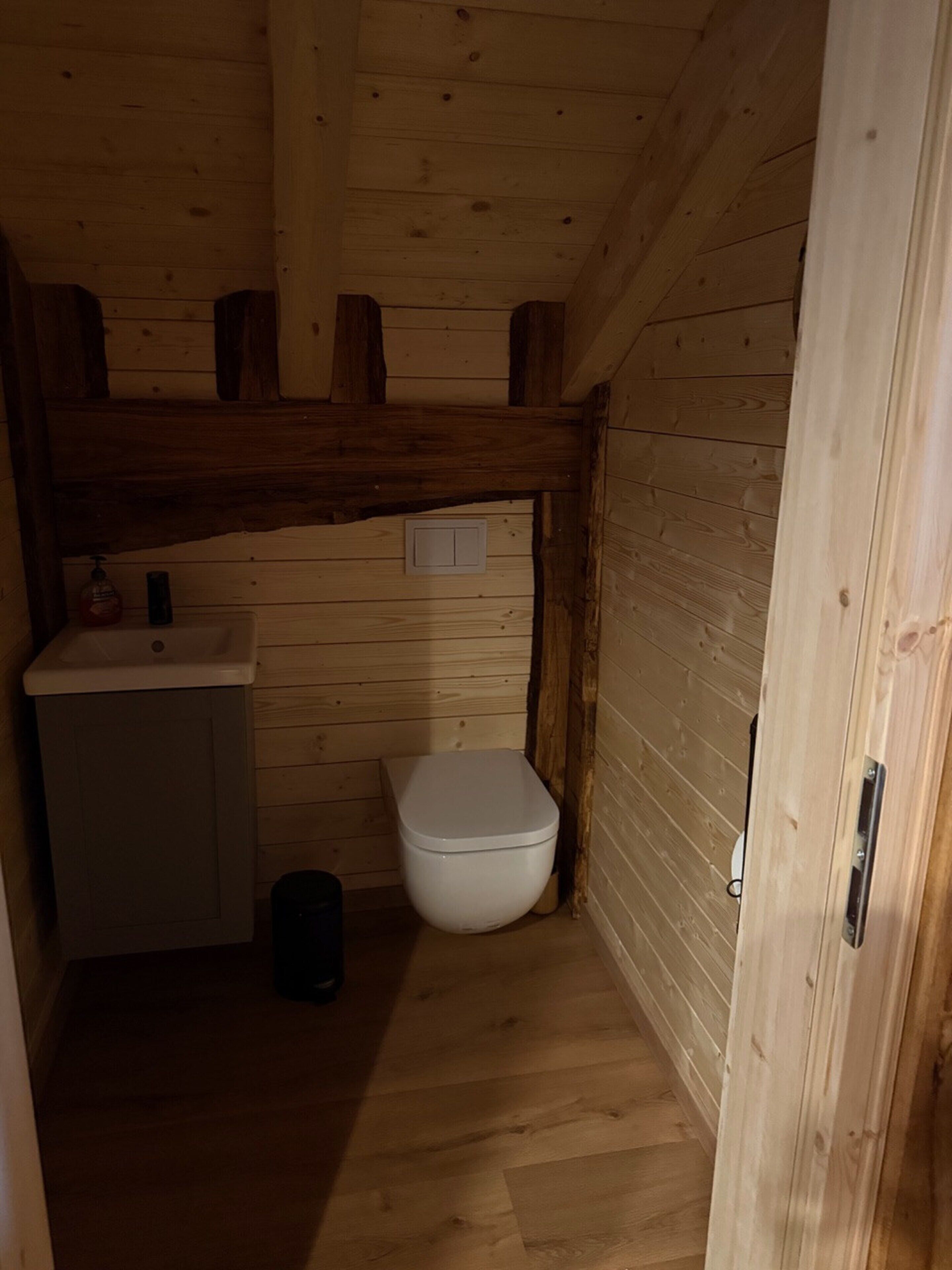 A small, wooden cabin bathroom with a toilet, sink, and a small trash bin.