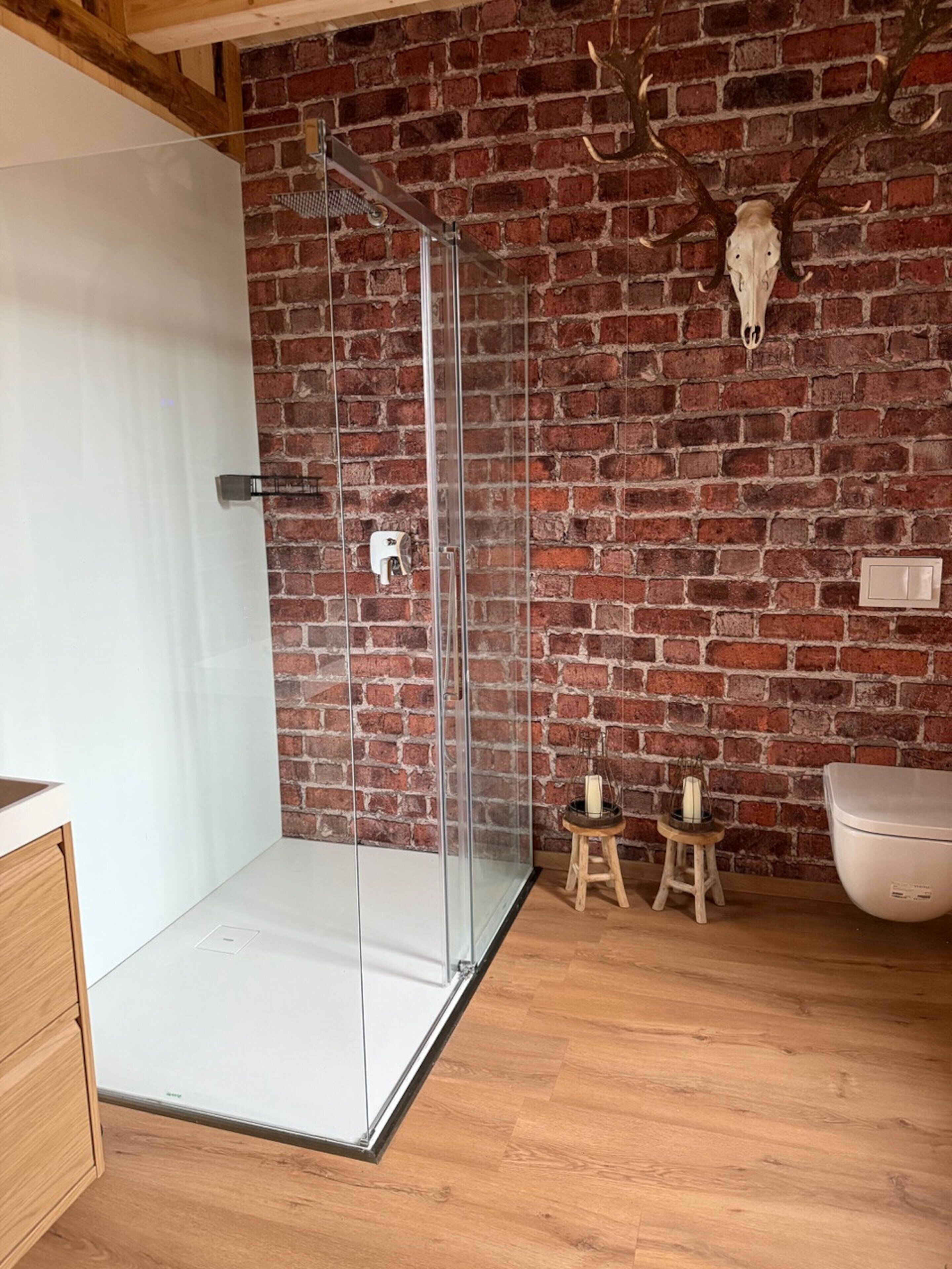 A modern bathroom with a glass shower enclosure, a brick wall, a wooden floor, and a rustic decor including a skull and candles.