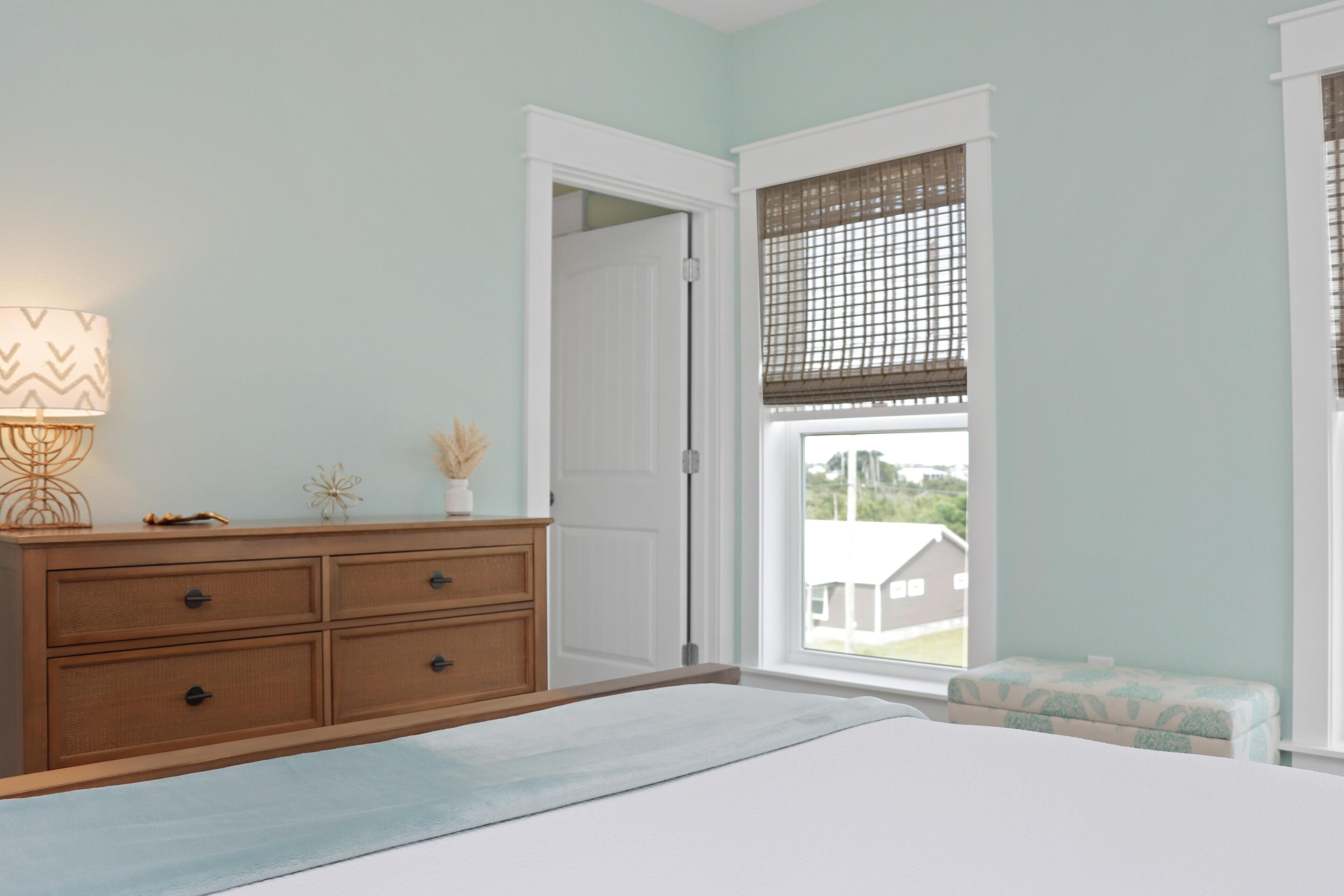 A bedroom with a bed, a dresser, a window with blinds, and a view of a house outside.