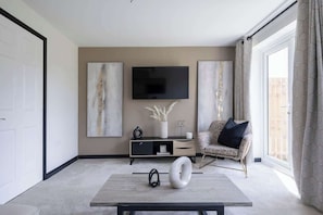 Una sala de estar moderna con un televisor de pantalla plana, arte abstracto en la pared, un sofá, una mesa de centro y un amplio ventanal con cortinas.