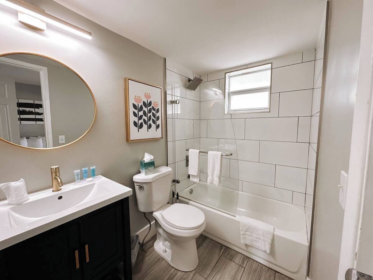 A bathroom with a white sink, toilet, and bathtub. There is a mirror with a gold frame, a framed picture on the wall, and a window above the bathtub.