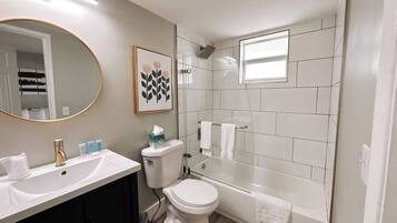 A bathroom with a white sink, toilet, and bathtub. There is a mirror with a gold frame, a framed picture on the wall, and a window above the bathtub.