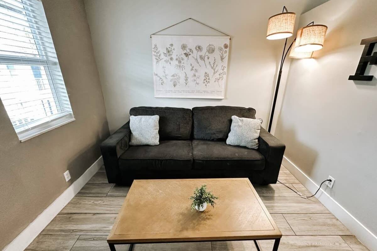 Ein Wohnzimmer mit einem dunkelbraunen Sofa, zwei hellen Kissen, einem Couchtisch aus Holz mit einer Pflanze, einem an der Wand befestigten Pflanzenmotiv und zwei Stehlampen.