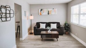 Ein Wohnzimmer mit einem dunklen Sofa, einem Couchtisch und einer Stehlampe. Rechts befindet sich ein Fenster mit Jalousien und an der Wand ein Gemälde.