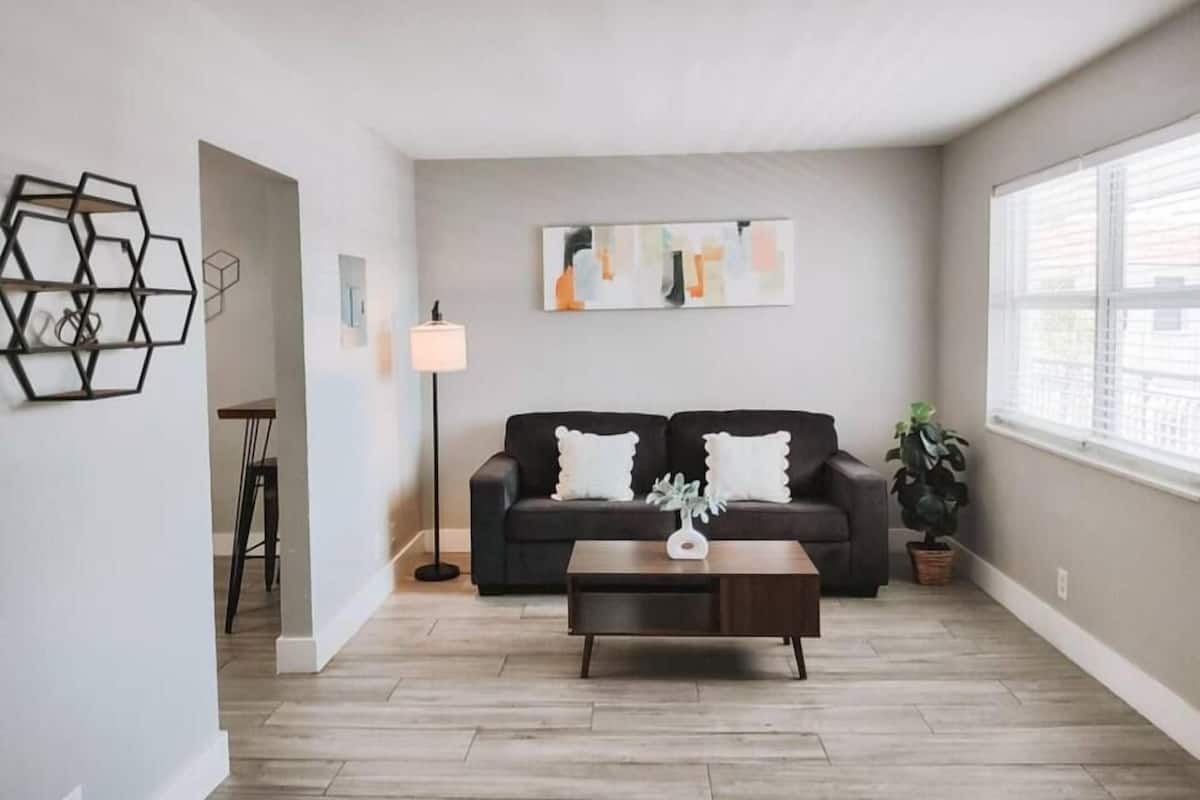 Ein Wohnzimmer mit einem dunklen Sofa, einem Couchtisch und einer Stehlampe. Rechts befindet sich ein Fenster mit Jalousien und an der Wand ein Gemälde.