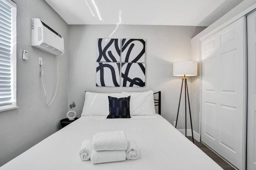 Un dormitorio moderno con una cama, aire acondicionado de pared, una mesita de noche con lámpara y una obra de arte abstracta de gran tamaño sobre la cama.
