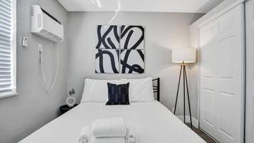 Un dormitorio moderno con una cama, aire acondicionado de pared, una mesita de noche con lámpara y una obra de arte abstracta de gran tamaño sobre la cama.
