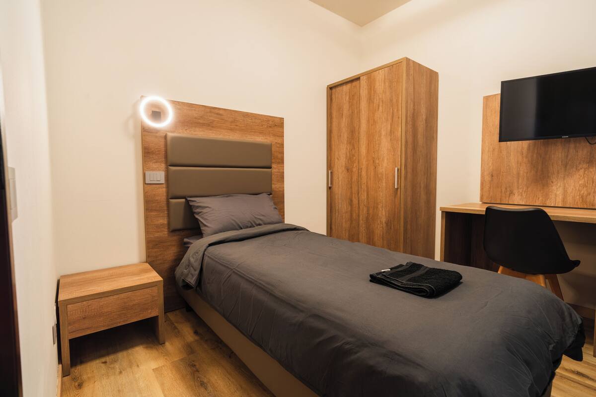 Une chambre à coucher comprenant un lit, une table de chevet, une armoire en bois, un bureau avec une chaise et un téléviseur fixé au mur.