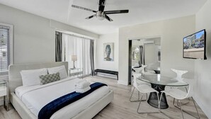 Een moderne hotelkamer met een bed, een eethoek met een glazen tafel en witte stoelen, een plafondventilator en uitzicht op de stad.