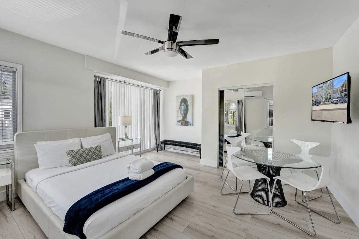 Una habitación de hotel moderna con una cama, un comedor con una mesa de vidrio y sillas blancas, un ventilador de techo y vistas a la ciudad.