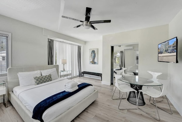 Quarto de hotel moderno com cama, área de jantar com mesa de vidro e cadeiras brancas, ventoinha no teto e vista para a cidade.
