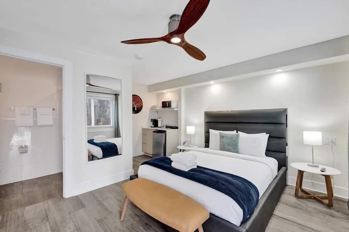Una habitación de hotel moderna con una cama grande, un ventilador de techo, una pequeña cocina americana y un baño visible a través de una puerta abierta.