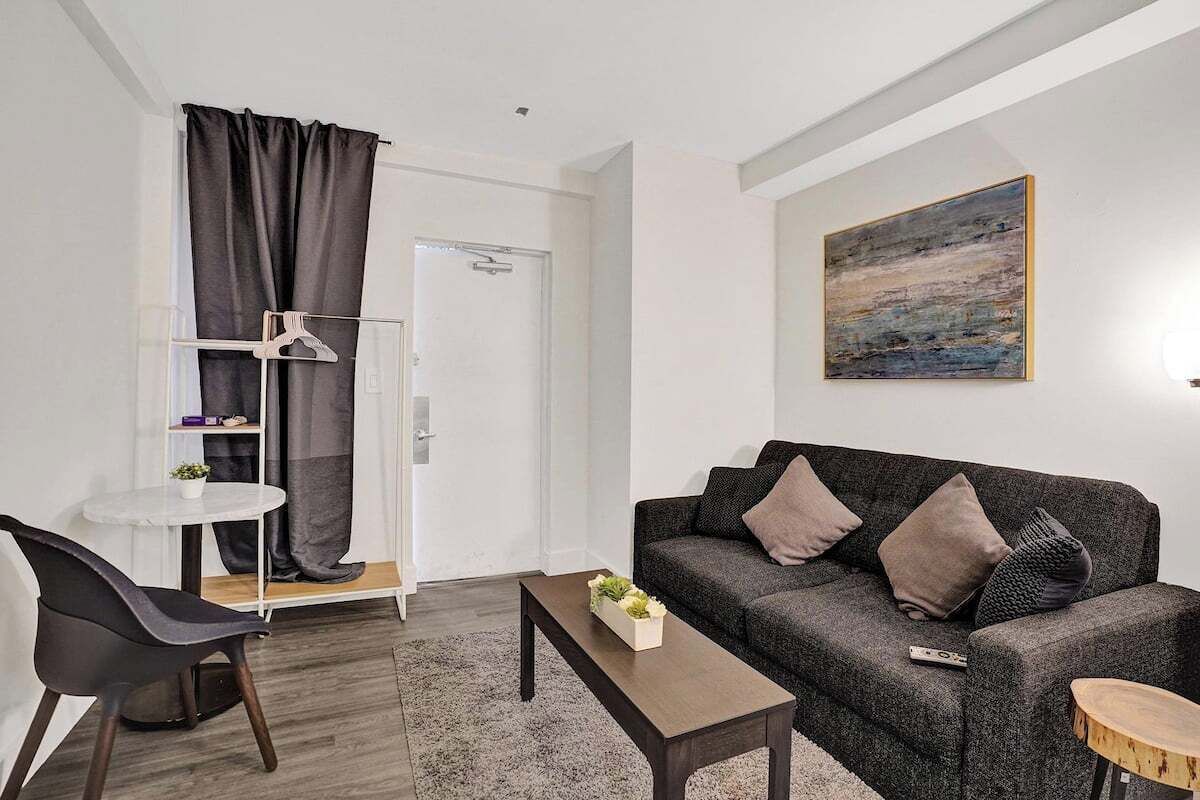 Una sala de estar moderna con un sofá gris, una pequeña mesa redonda y una silla negra. Hay una puerta blanca con perchero y un cuadro en la pared.