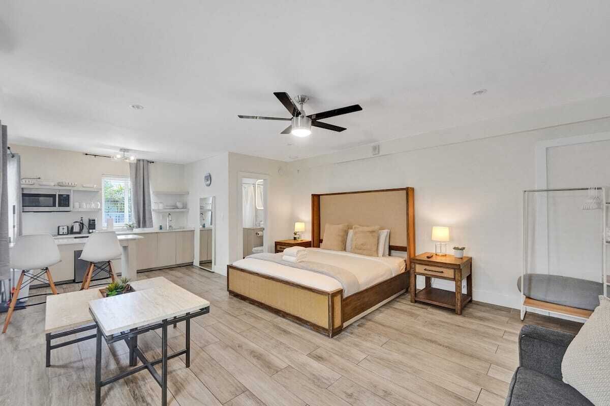 A modern hotel room with a large bed, a dining table, a kitchenette, and a separate seating area.