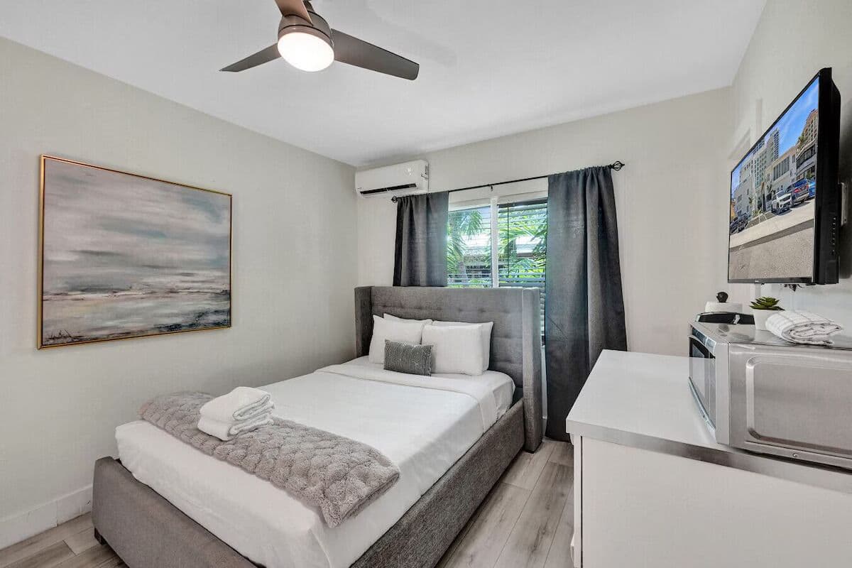 Un dormitorio moderno con cama, ventilador de techo, televisor de pantalla plana y ventana con cortinas.