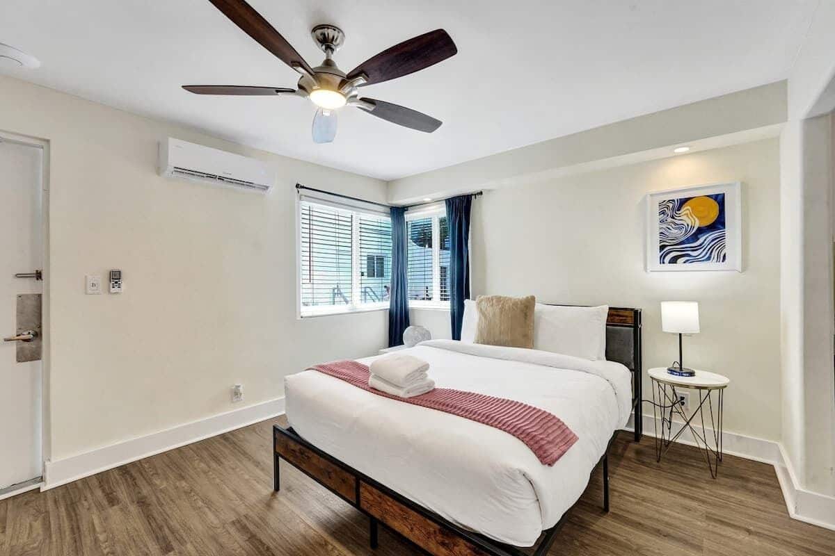 Habitación de hotel con cama, ventilador de techo, aire acondicionado, ventana con persianas, un cuadro en la pared y una mesita con lámpara.