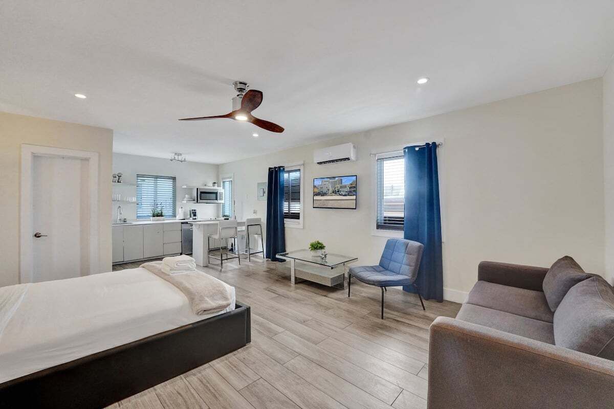 Une chambre d’hôtel moderne avec un lit, un canapé, une petite table, une chaise, un ventilateur de plafond et un coin cuisine à l’arrière-plan.