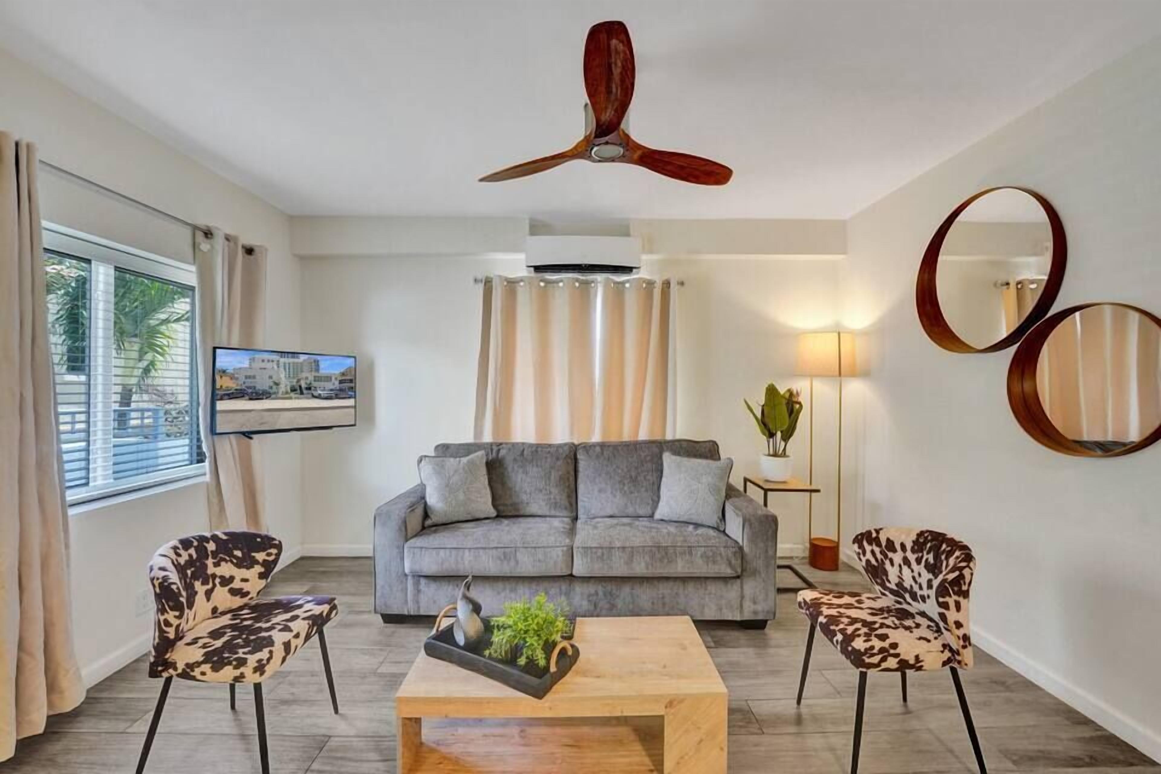 A modern living room with a grey sofa, two patterned armchairs, a wooden coffee table with a plant, a ceiling fan, and a TV on the wall.