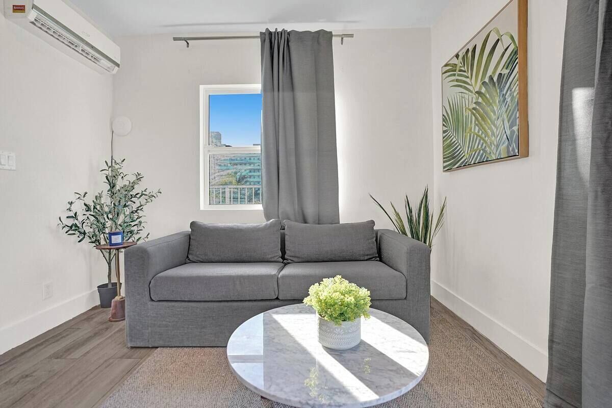Ein modernes Wohnzimmer mit grauem Sofa, einem runden Couchtisch mit Pflanze, einem Fenster mit Blick auf Gebäude und einer großen Pflanze rechts.