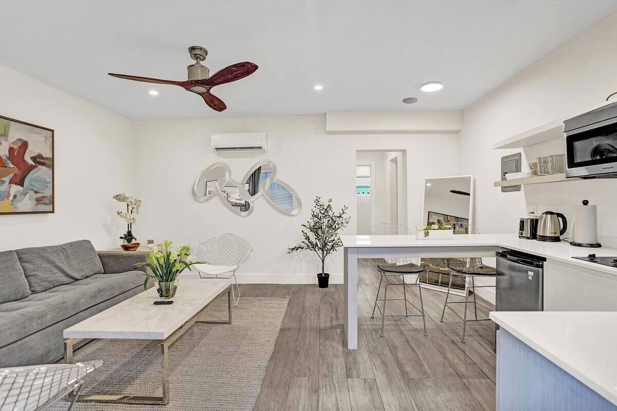 A modern kitchen with a central island, a dining area with a white table and chairs, a grey sofa, and a ceiling fan.