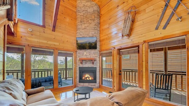 A cozy cabin interior with a stone fireplace, leather sofas, and large windows offering a view of the outdoors. - Rustic Retreat – Jacuzzi, Fireplaces & Views (Sevierville)