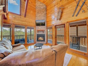 A cozy cabin interior with a stone fireplace, leather sofas, and large windows offering a view of the outdoors.