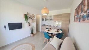 Ein modernes Wohnzimmer mit einer Couch, einem Esstisch mit Stühlen, einem an der Wand montierten Fernseher und einer Küchenzeile mit einer Pendelleuchte.