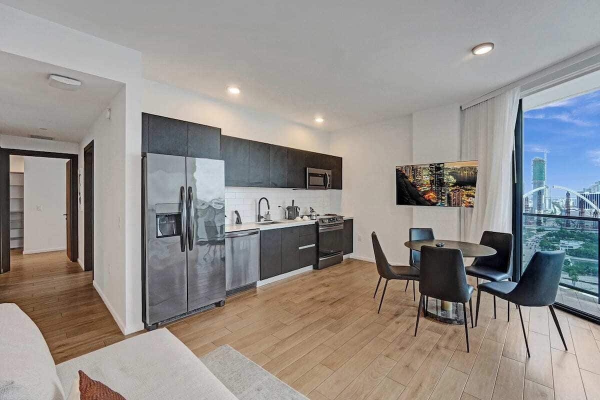 Una cocina moderna con refrigerador, microondas y estufa. Hay un comedor con mesa y sillas. Un ventanal ofrece vistas a un paisaje urbano.