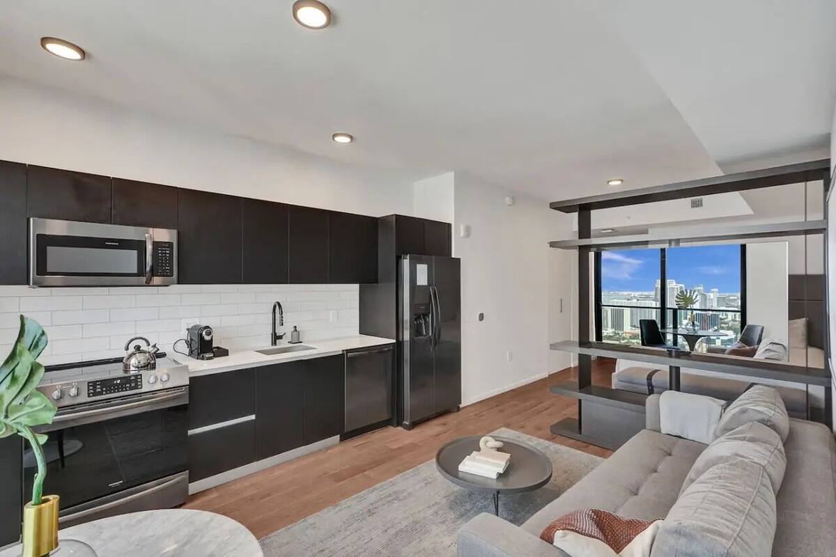 Una cocina moderna con armarios oscuros, un refrigerador de acero inoxidable y un microondas. Hay un área de comedor con un sofá y una mesa, y vistas de la ciudad a través de amplias ventanas.