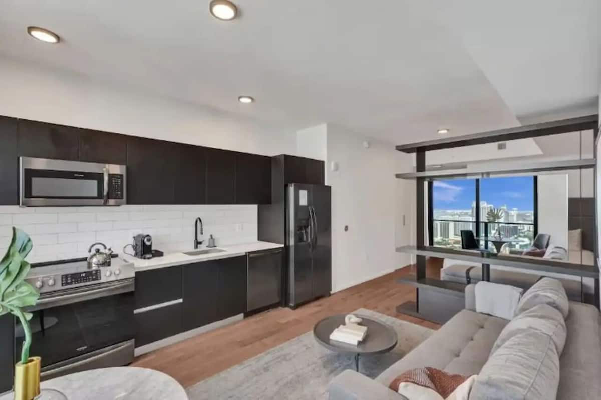 Una cocina moderna con armarios oscuros, un microondas de acero inoxidable y un refrigerador. Hay un comedor con un sofá y una mesa de centro. Un ventanal ofrece vistas a la ciudad.