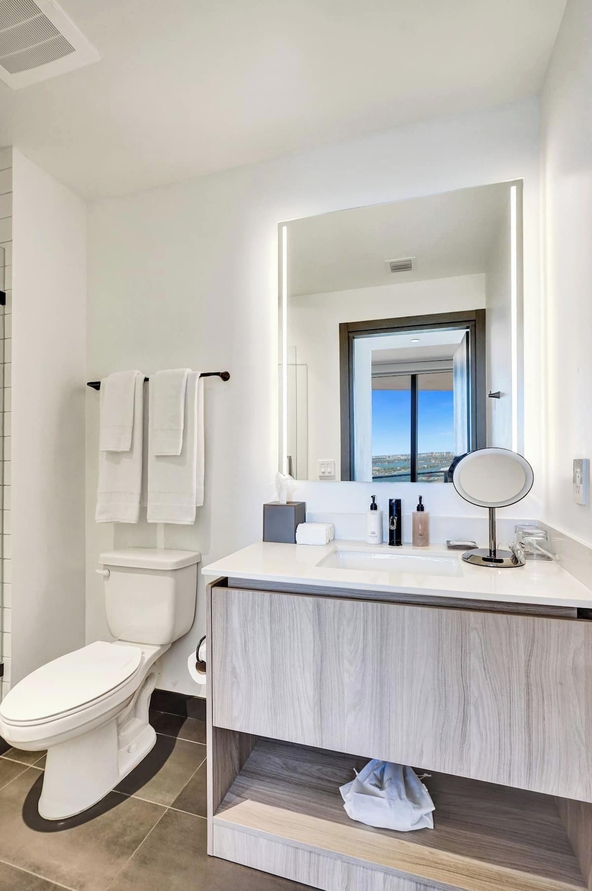 Une salle de bain moderne équipée de toilettes, d’un meuble-lavabo avec miroir et offrant une vue sur la ville par la fenêtre.
